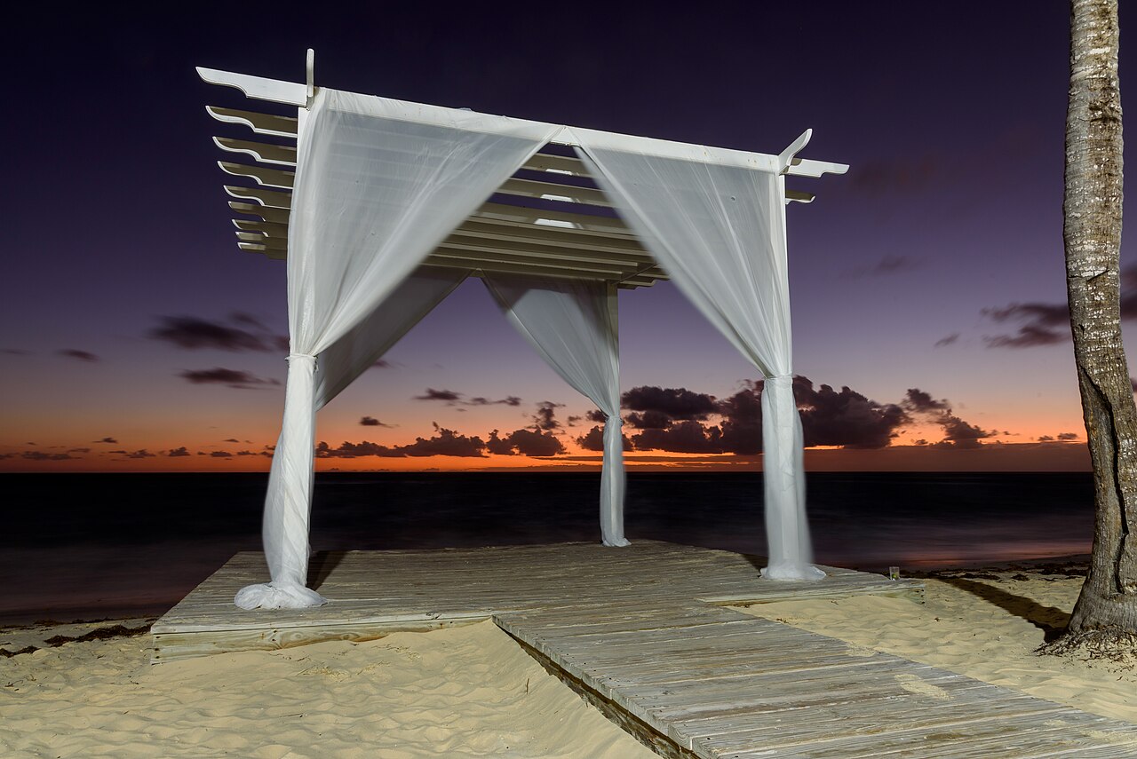 A wedding canopy draped in white fabric on a beach boardwalk at dusk