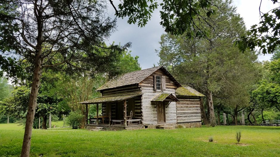 A historic log cabin with a porch under tall trees on a green lawn
