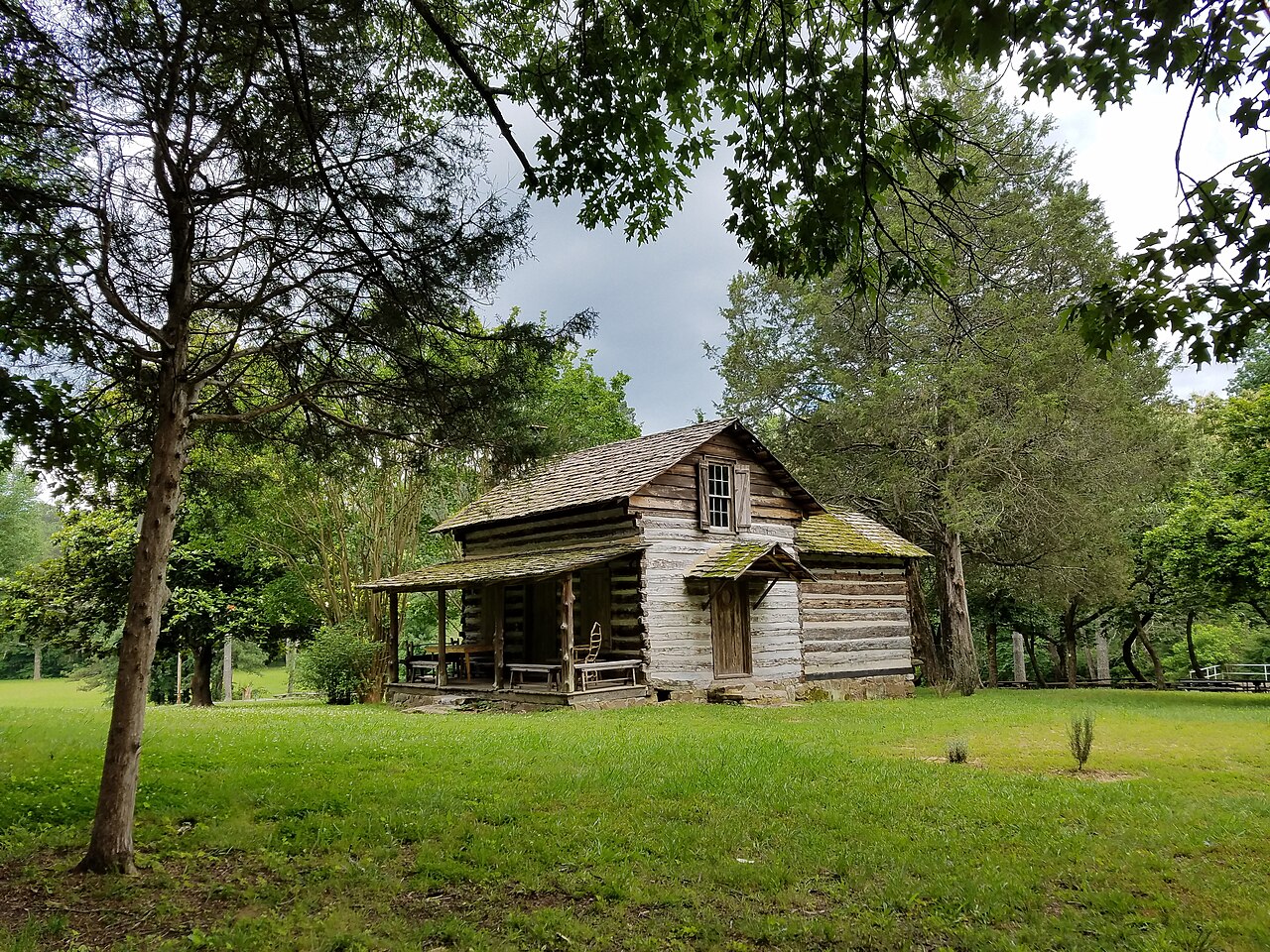 A historic log cabin with a porch under tall trees on a green lawn